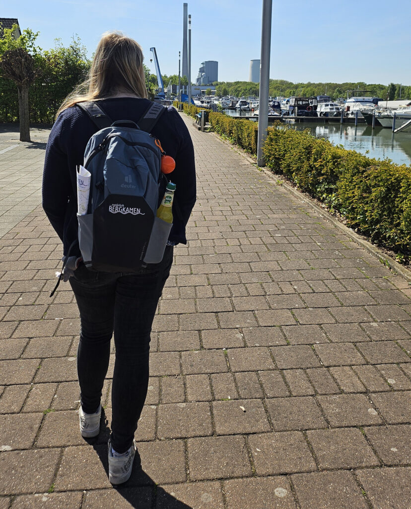 Rückenansicht einer Wanderin mit einem Rucksack, der das Logo der Stadt Bergkamen trägt, beim Gehen entlang der Promenade der Marina Rünthe mit Booten im Hintergrund.
