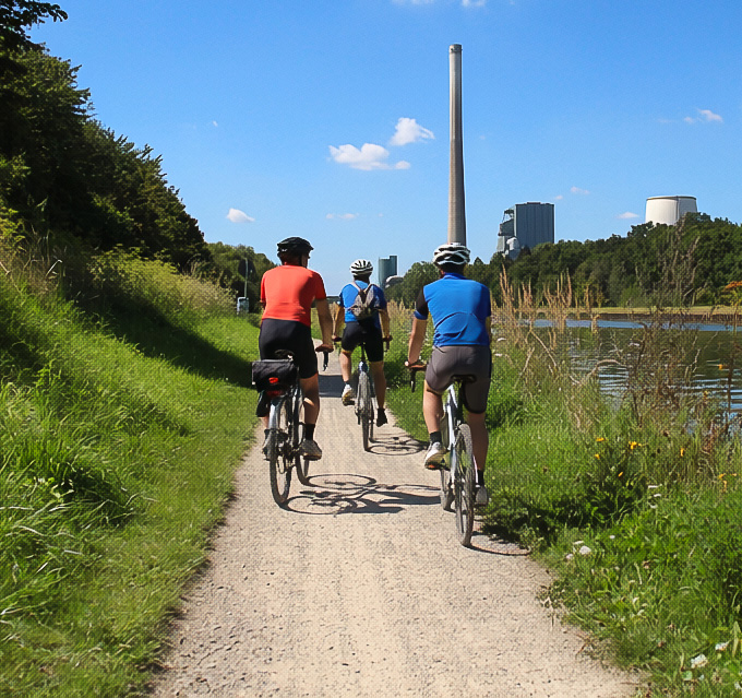 Drei Radfahrer auf dem Weg entlang des Datteln-Hamm-Kanals mit grüner Uferkulisse und dem Kraftwerk Bergkamen im Hintergrund.