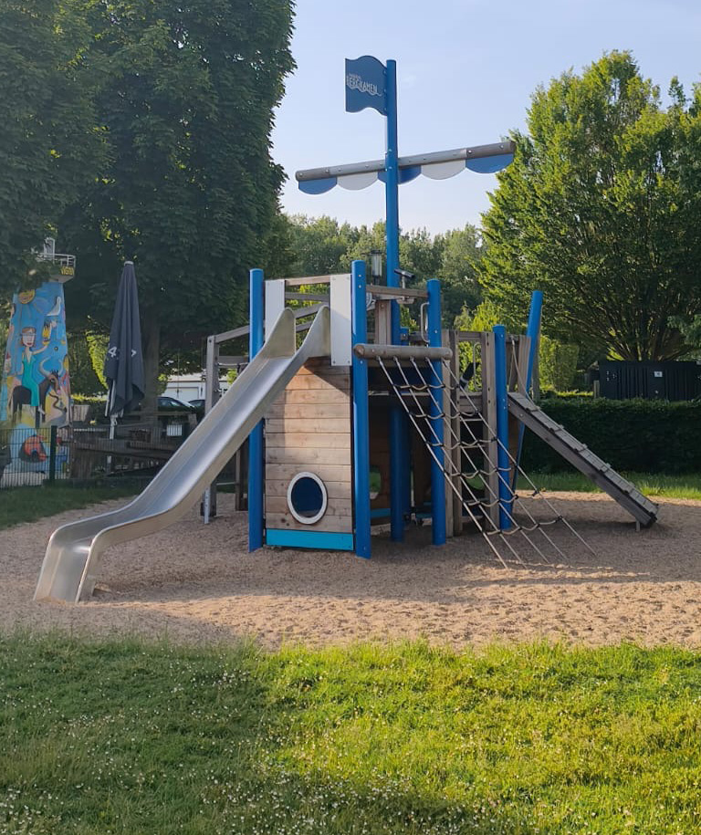 Kletter-Piratenboot auf einem Sandspielplatz, umgeben von einer Grünfläche und Bäumen.